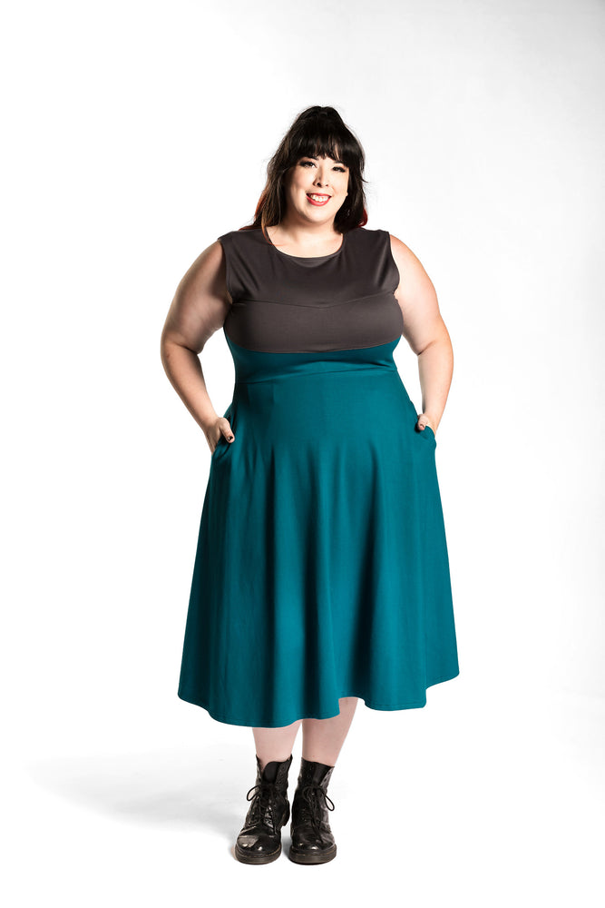 Katie Lynn is wearing the Fighter Longsword Dress - Teal. Both hands are in her skirt pockets. Katie Lynn is wearing a 2X. Her measurements are a 49" Bust, 40" Waist, and 53" Hips, and she is 5'8"